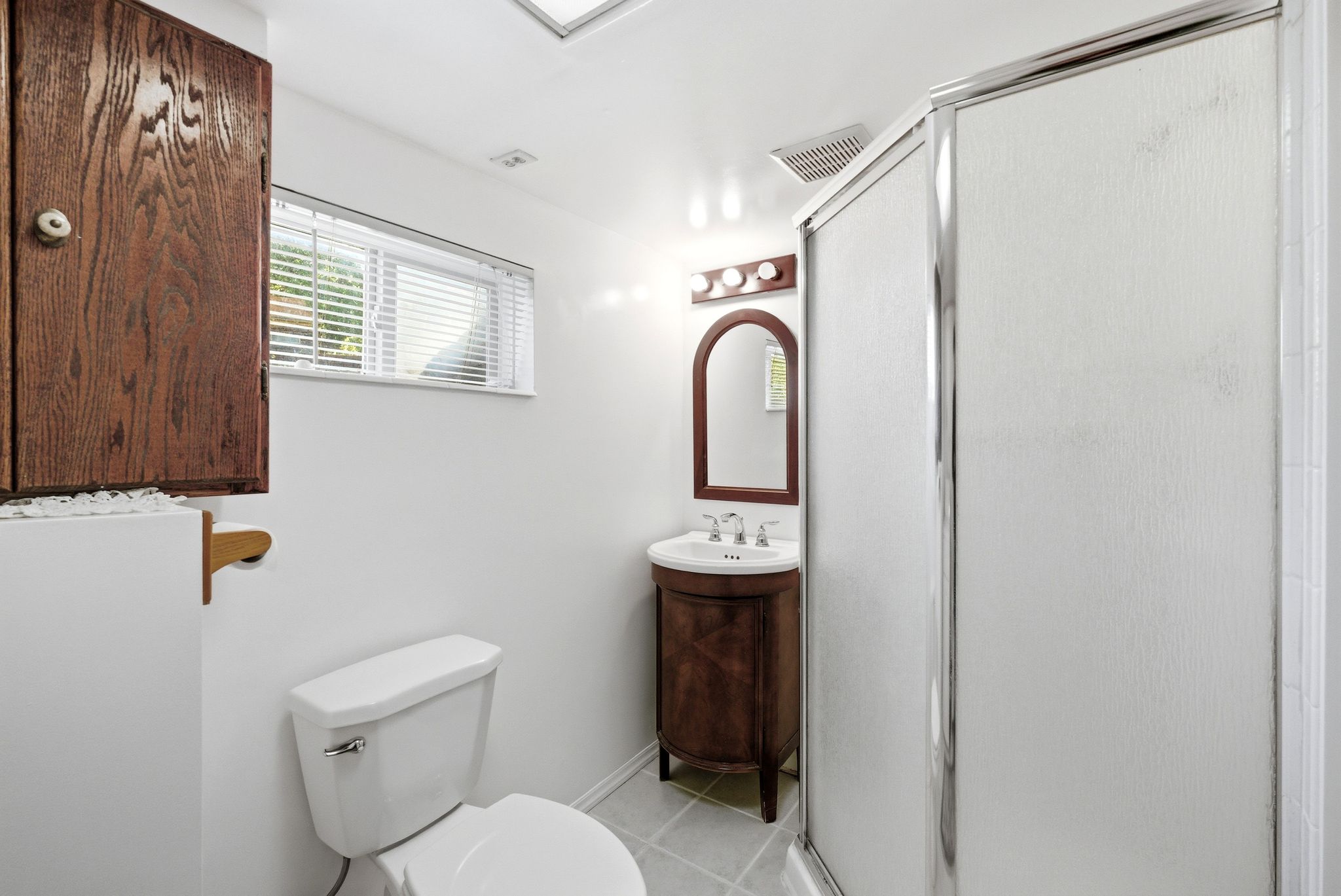 Bathroom featuring a glass-enclosed shower, a wood-finish vanity with an arched mirror, an overhead light fixture, a window with blinds, and white square floor tiles