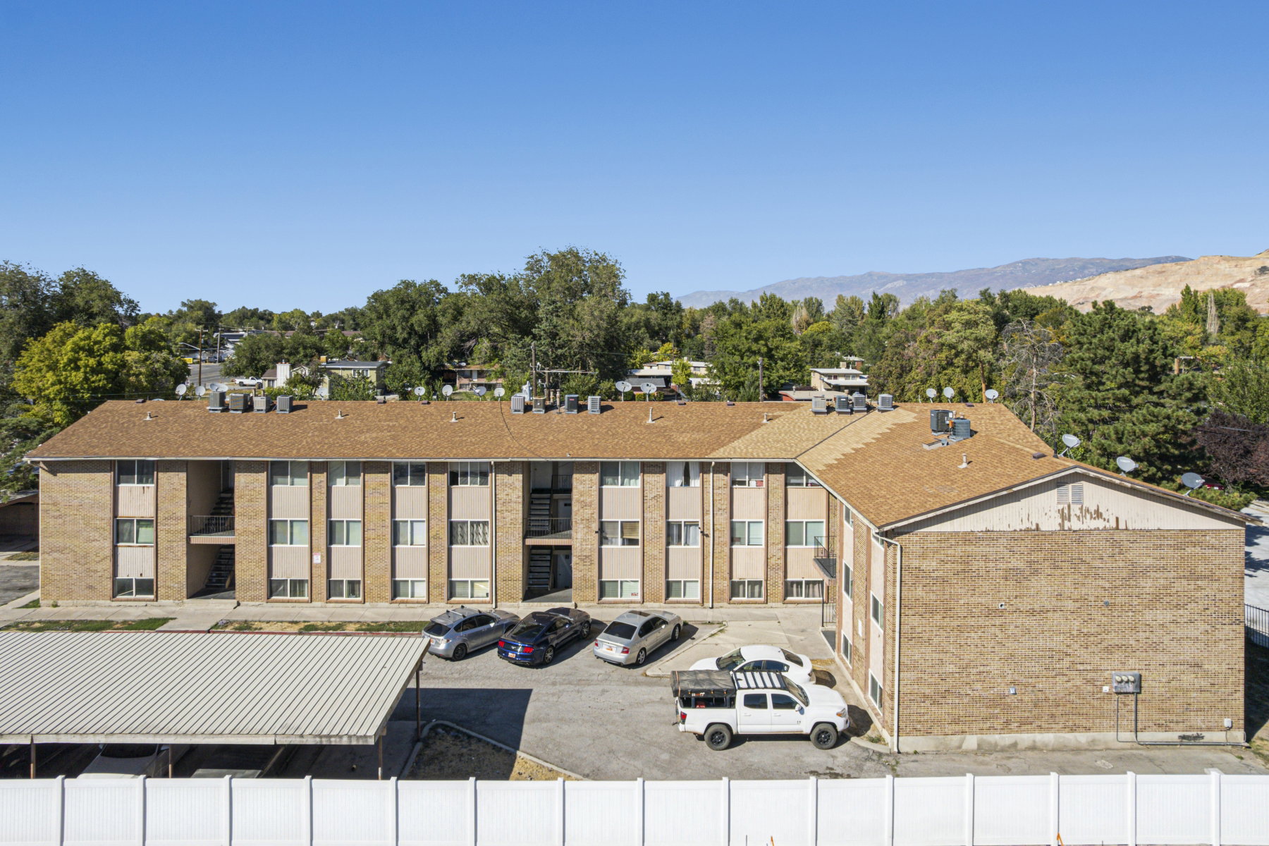 Multi-story residential building featuring brick and panel facades, brown shingle roofing, surface parking, and a covered parking structure