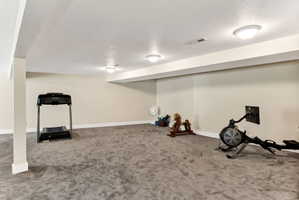 Spacious lower-level room featuring light neutral wall tones, gray carpeting, recessed ceiling lighting, and exposed structural beams