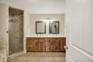 Spacious primary bathroom featuring a glass-enclosed shower with stone-look tile surround, a double vanity with distressed wood-finish cabinetry, a white countertop, and wood-finish tile flooring