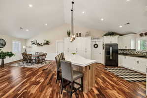 Open-concept kitchen and dining area featuring vaulted ceilings, wood-finish flooring, white cabinetry, a kitchen island with seating, and a black refrigerator