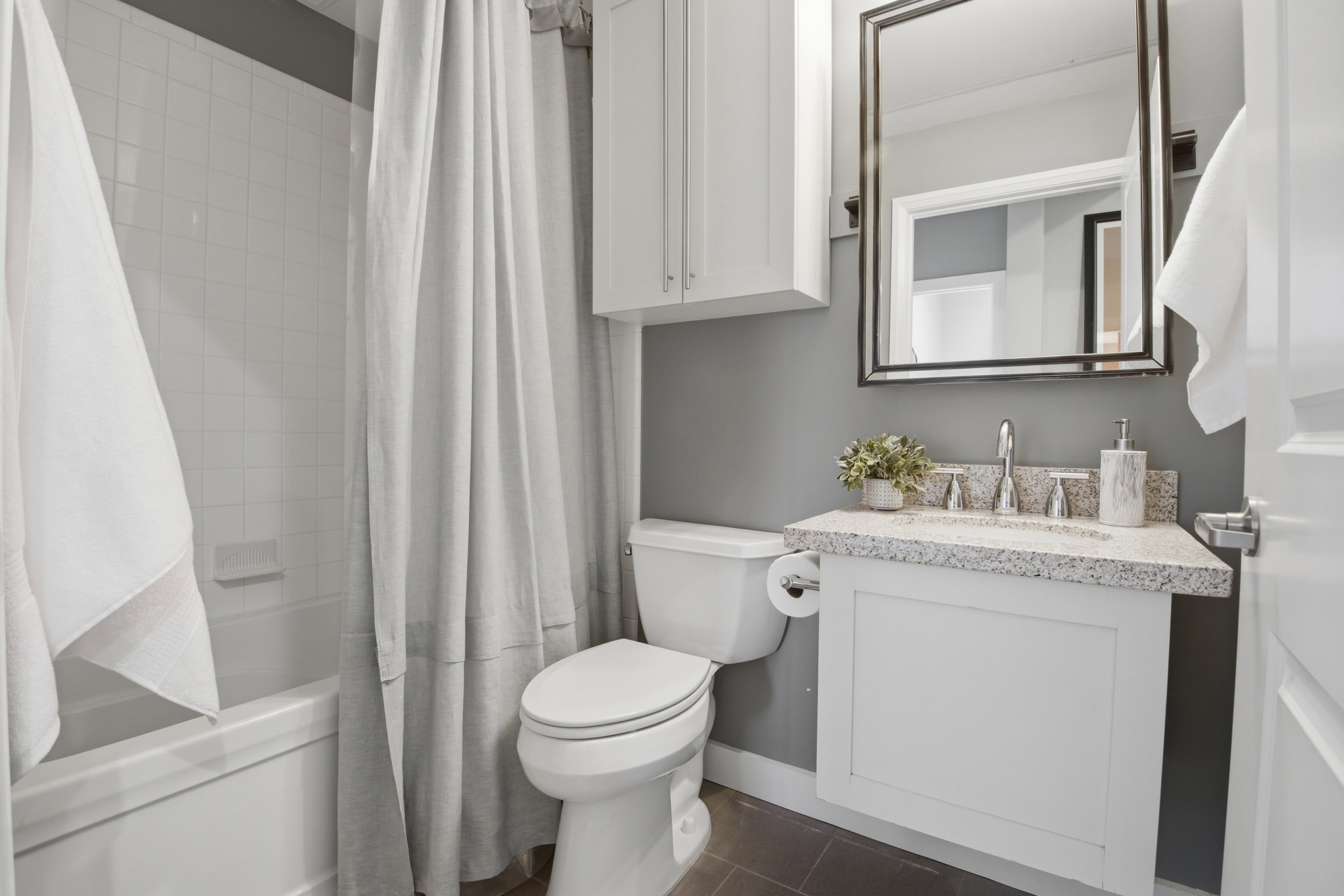 Bathroom featuring a white subway tile bath/shower surround, a white vanity with a speckled countertop, a rectangular framed mirror, and a white wall-mounted cabinet