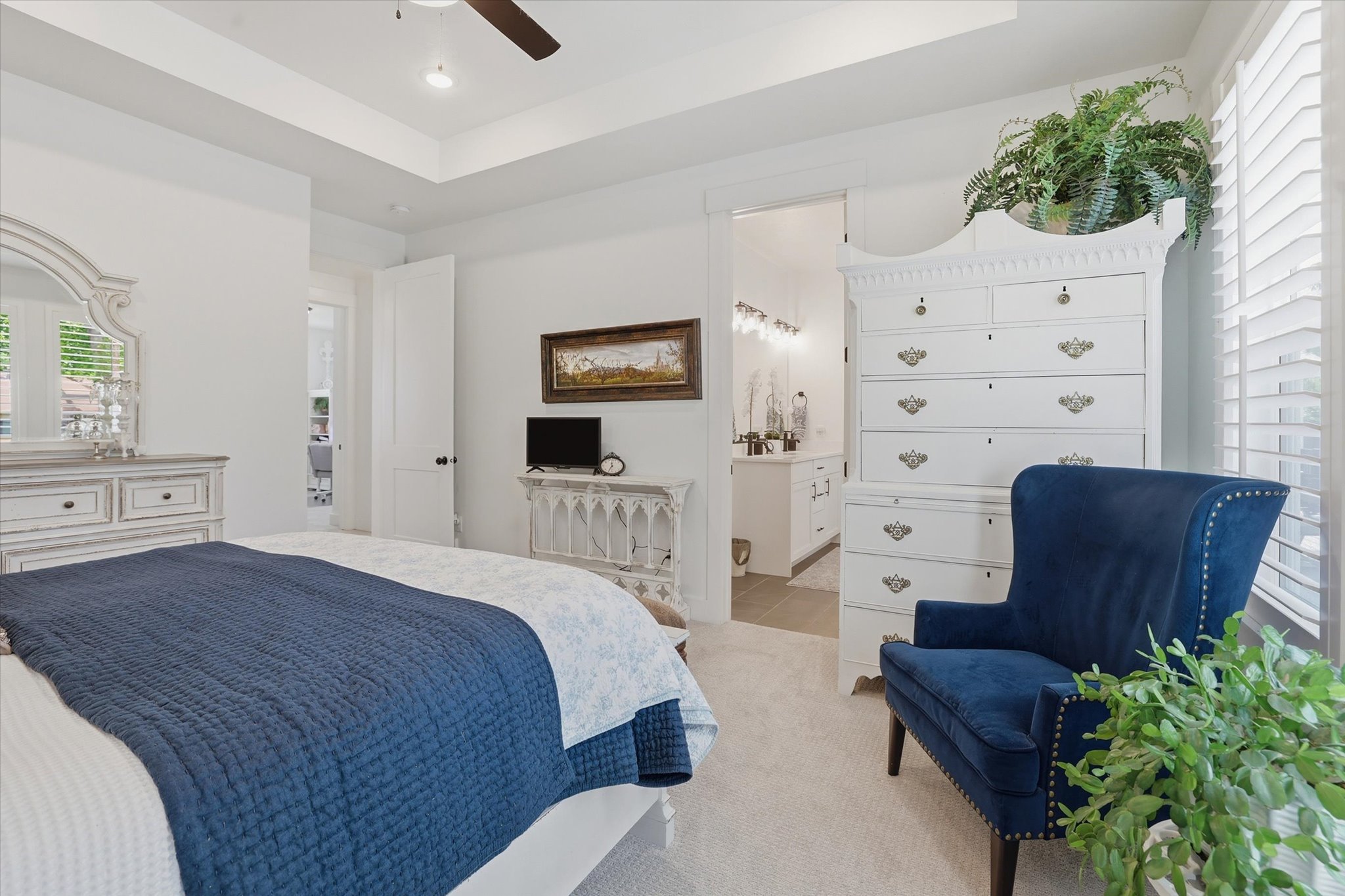 Carpeted room featuring a ceiling fan, tray ceiling, white plantation shutters, and an open doorway revealing a bathroom with a vanity and wall-mounted lighting