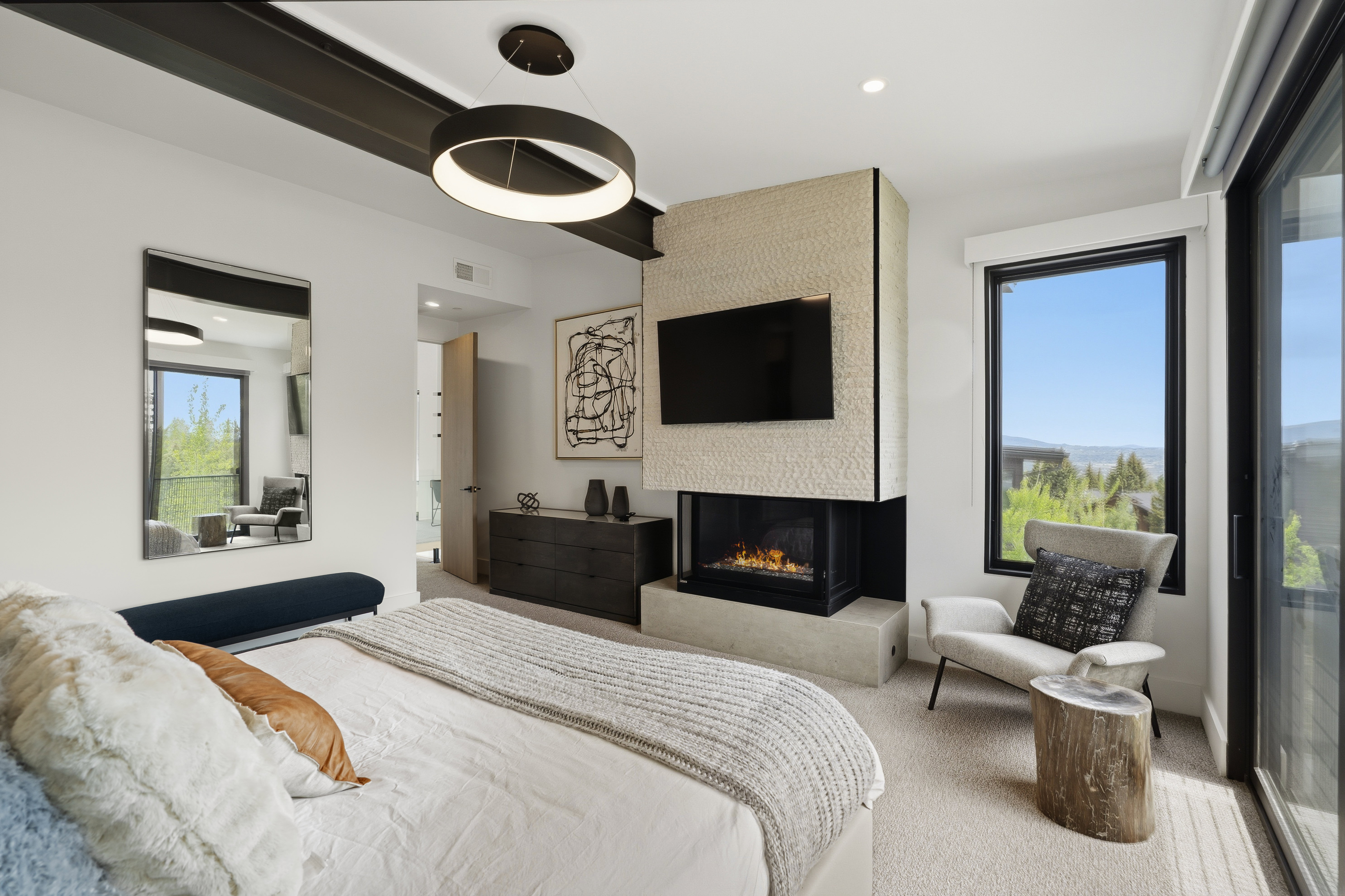 Modern bedroom featuring a stone-clad fireplace, integrated television, and a large window