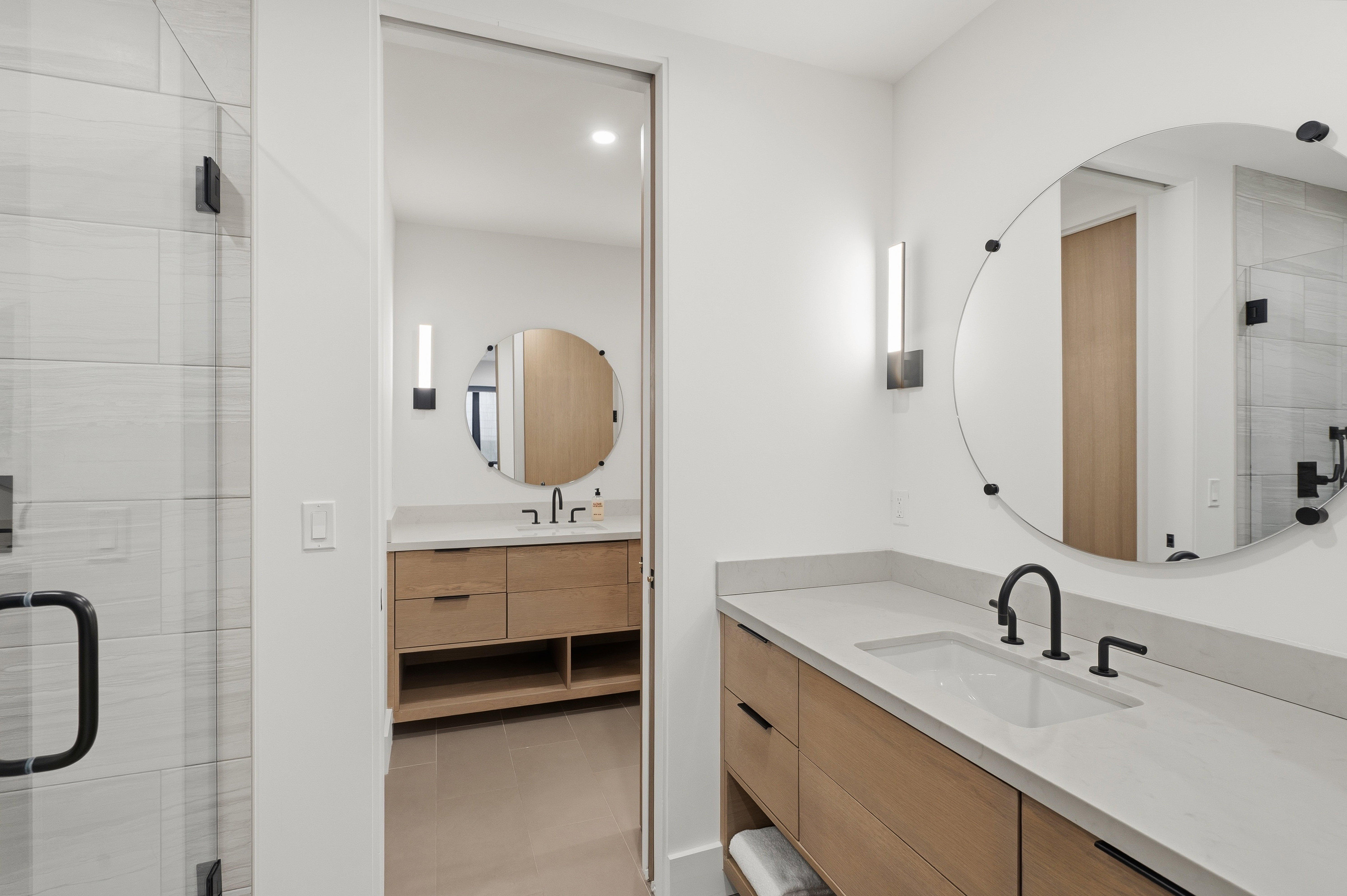 Contemporary bathroom featuring light wood-finish dual vanities with an integrated sink, matte black fixtures, and a large circular mirror