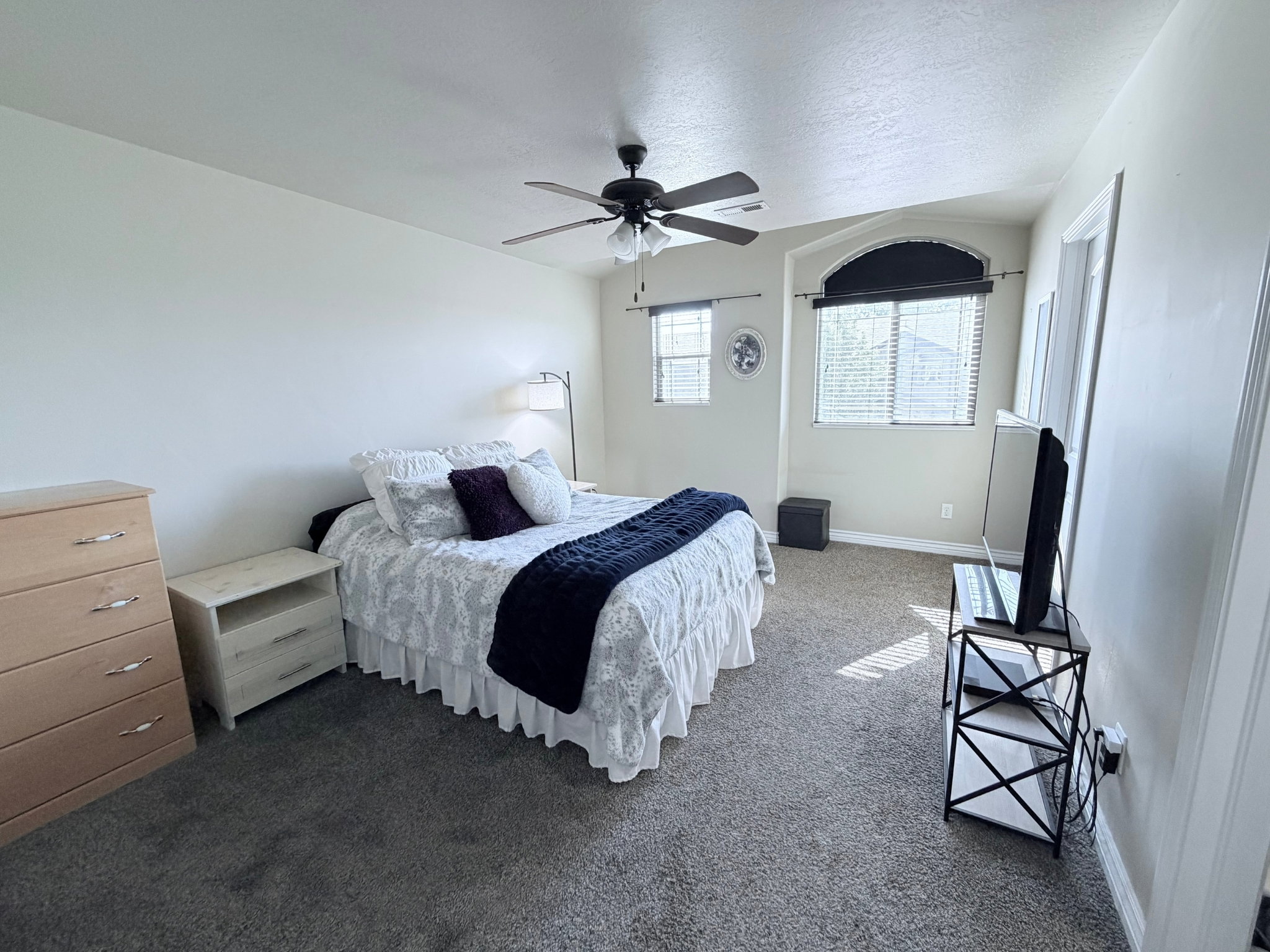 Spacious bedroom featuring light-toned walls with neutral carpeting, a ceiling fan with a light fixture, and multiple windows including an arched window with blinds