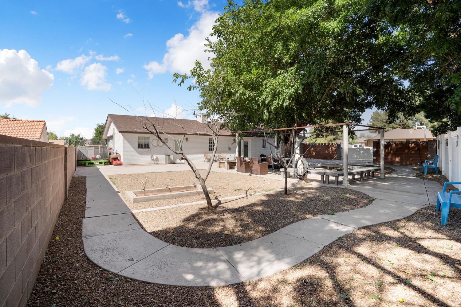 Spacious backyard featuring a concrete pathway, mature trees providing shade, a built-in outdoor structure, and a perimeter block wall