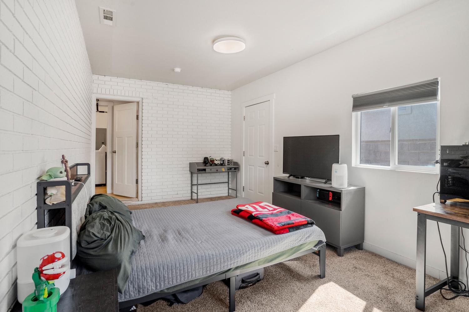 Carpeted bedroom featuring a white painted brick accent wall, a single window with blinds, and a flush-mount ceiling light