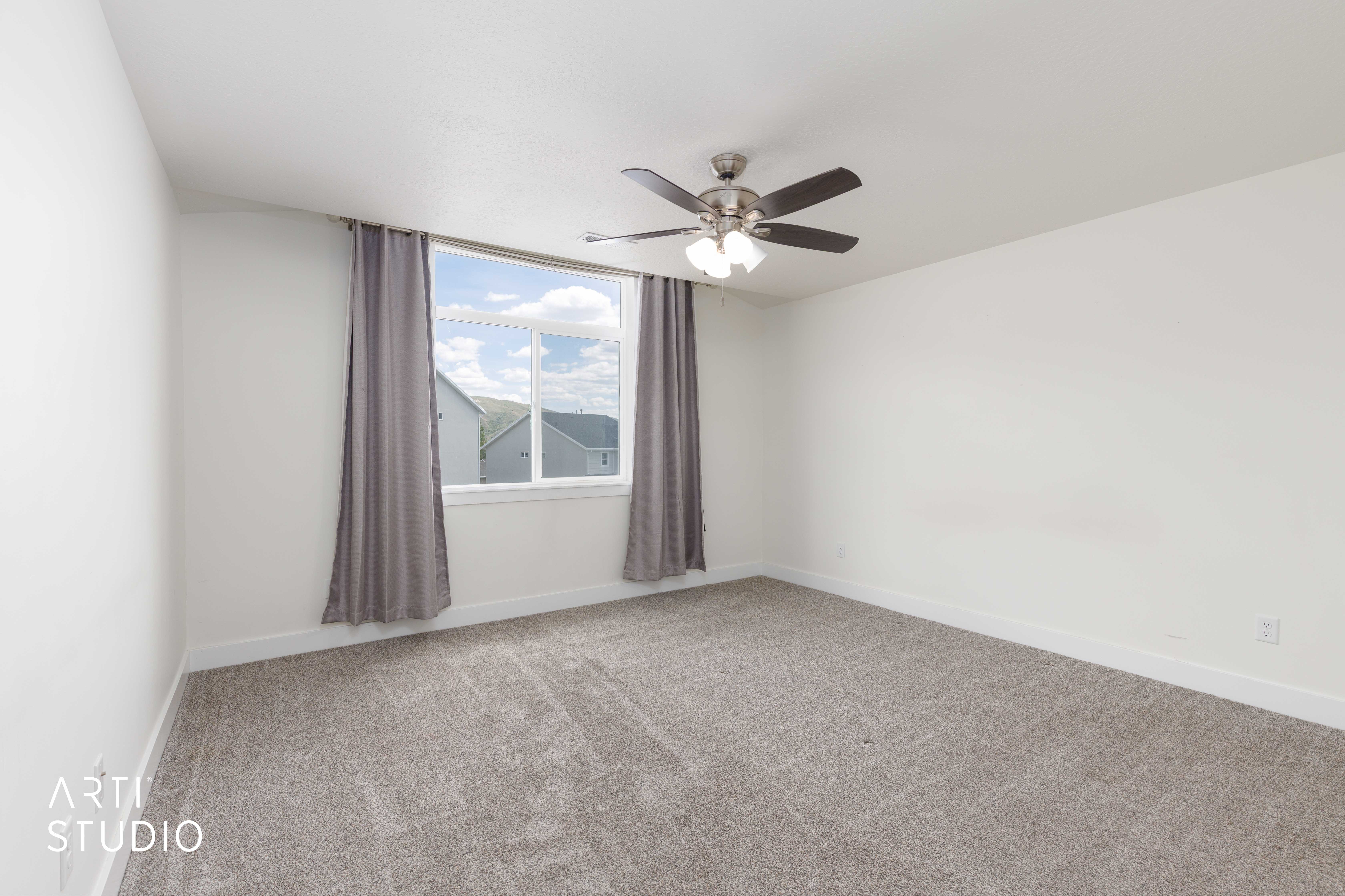 Spacious interior room featuring white walls, grey carpeting, a ceiling fan with integrated lighting, and a large window with grey drapes