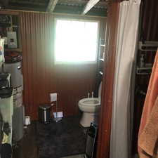 Bathroom featuring corrugated metal walls, a white framed window, a standard white toilet, wood-finish flooring, and a stand-up shower with a white curtain