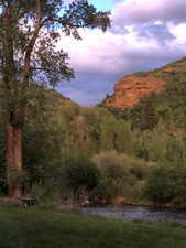 Expansive natural landscape featuring a flowing river, mature trees, verdant foliage, and distant red rock formations
