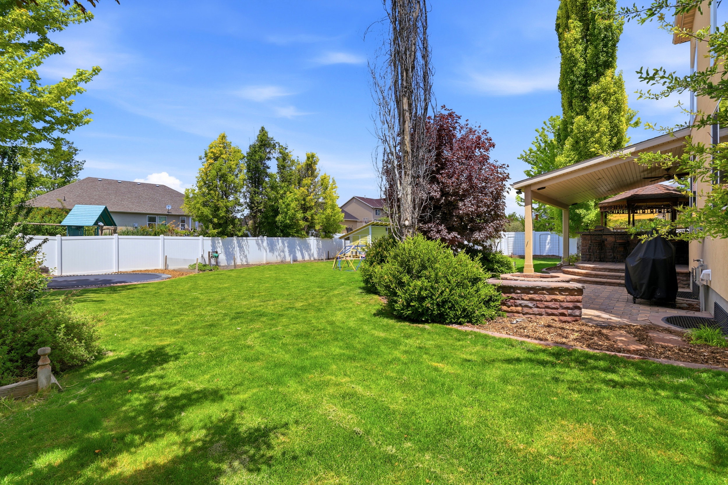 Expansive lawn area featuring a white privacy fence, mature trees, a covered patio, and a built-in brick fire pit