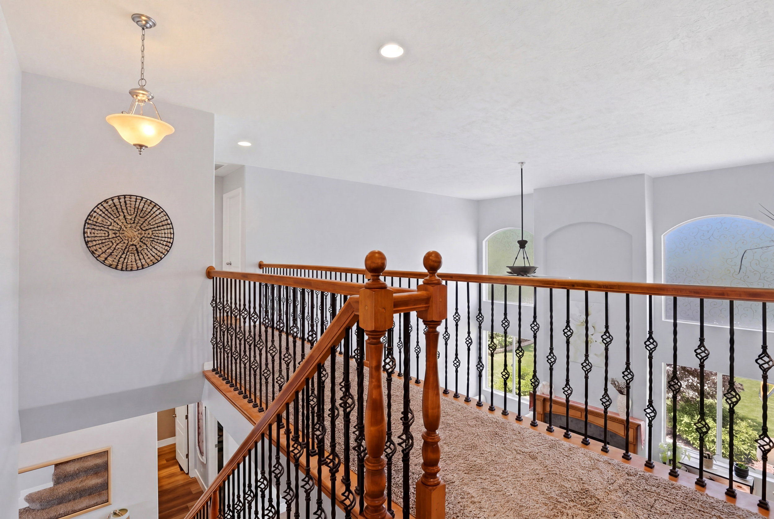 Upper level landing featuring light wood railings with decorative metal balusters, recessed lighting, and a neutral carpeted floor