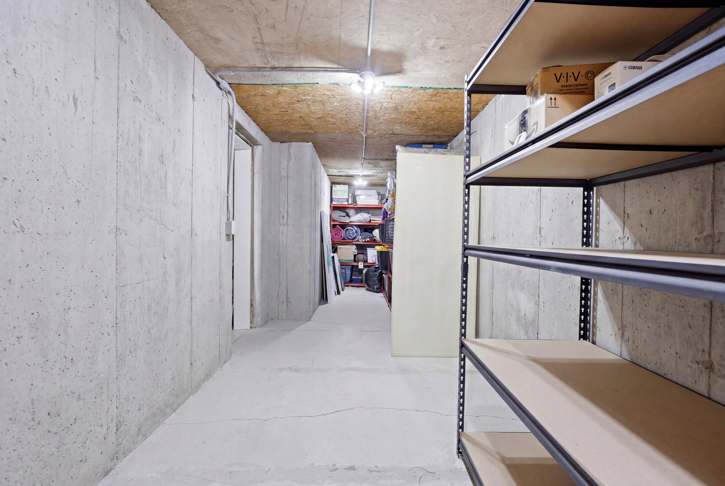 Unfinished basement area featuring concrete walls and flooring, exposed ceiling joists, and built-in metal shelving units