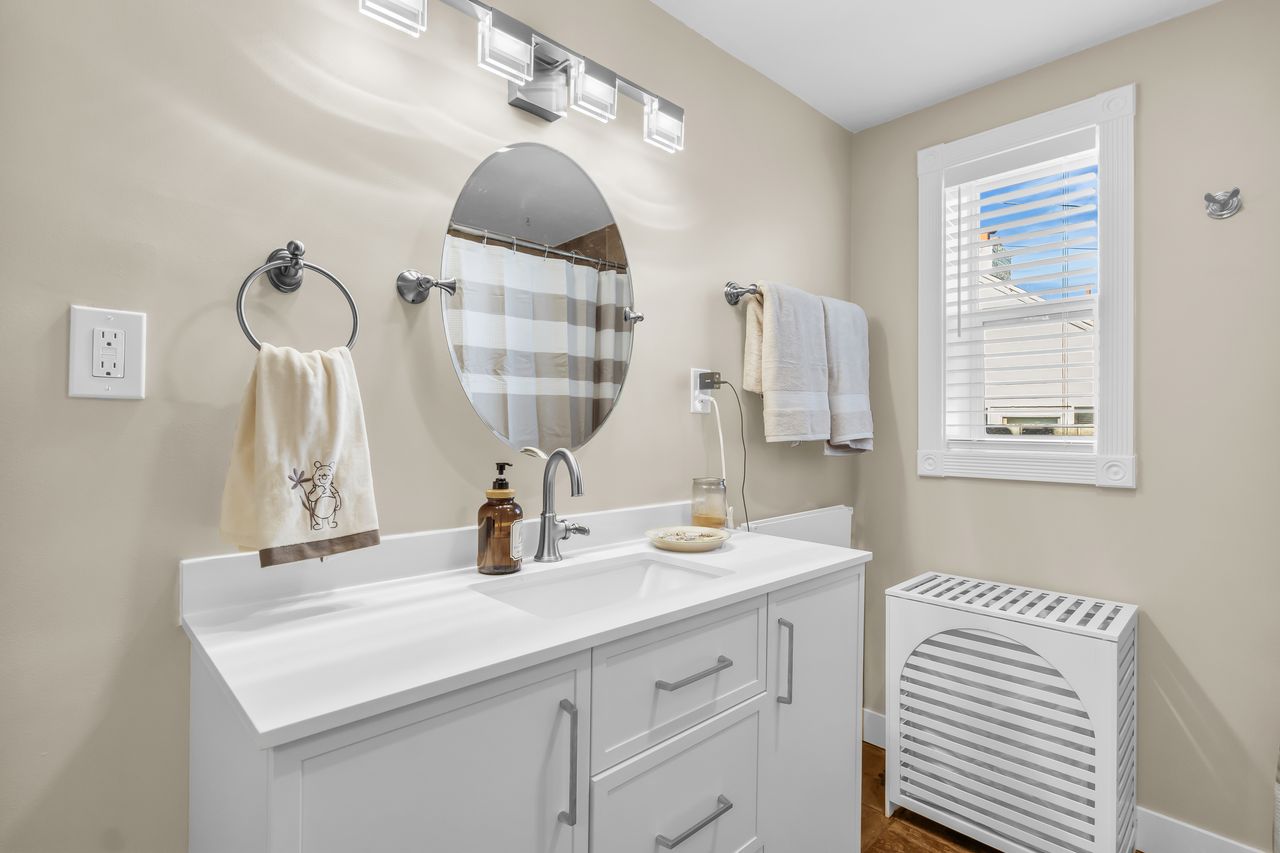 Contemporary bathroom featuring a white vanity with a solid surface countertop, an oval mirror, and a window with white blinds