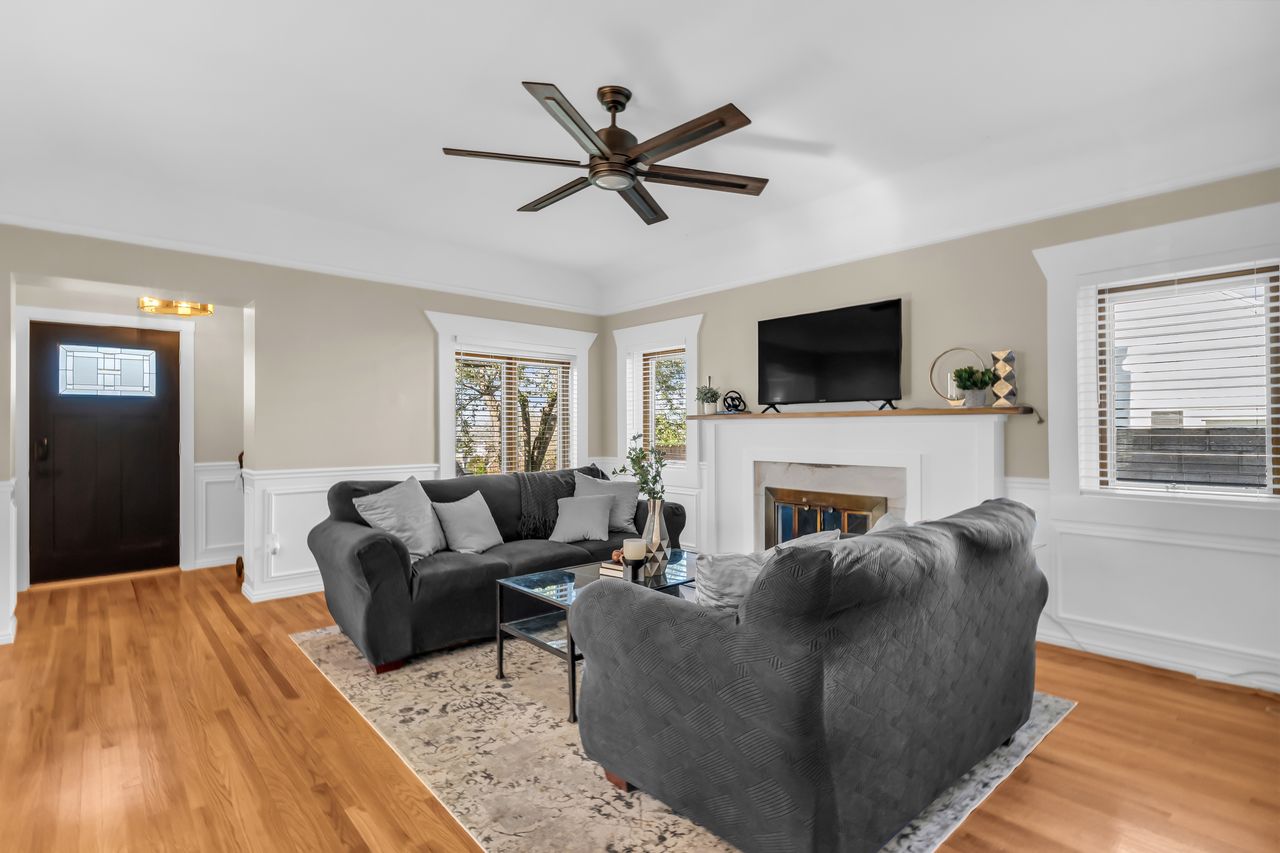 Hardwood flooring throughout featuring a fireplace with white mantel, a ceiling fan, and decorative wainscoting