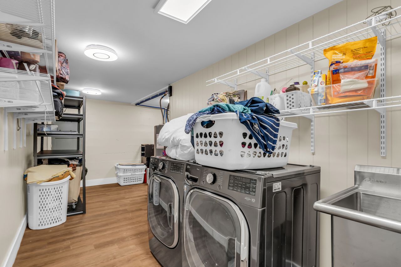 Dedicated laundry space featuring wood-finish flooring, two large capacity laundry appliances, a stainless steel utility sink, and extensive wall-mounted shelving