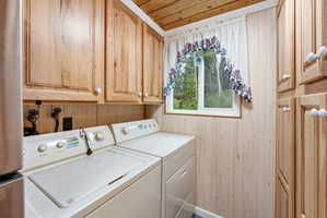 Utility room featuring light wood paneling, upper wood cabinetry, a window with exterior tree scenery, and a wood-finish ceiling