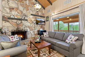 Stone fireplace with a black metal screen, wood-finish flooring, wood-paneled walls, vaulted ceiling with exposed wood beams, and a wide window overlooking trees