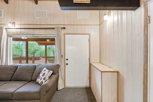 Interior space featuring light wood paneling, dark exposed ceiling beams, and a large window overlooking a wooden deck and lush green trees