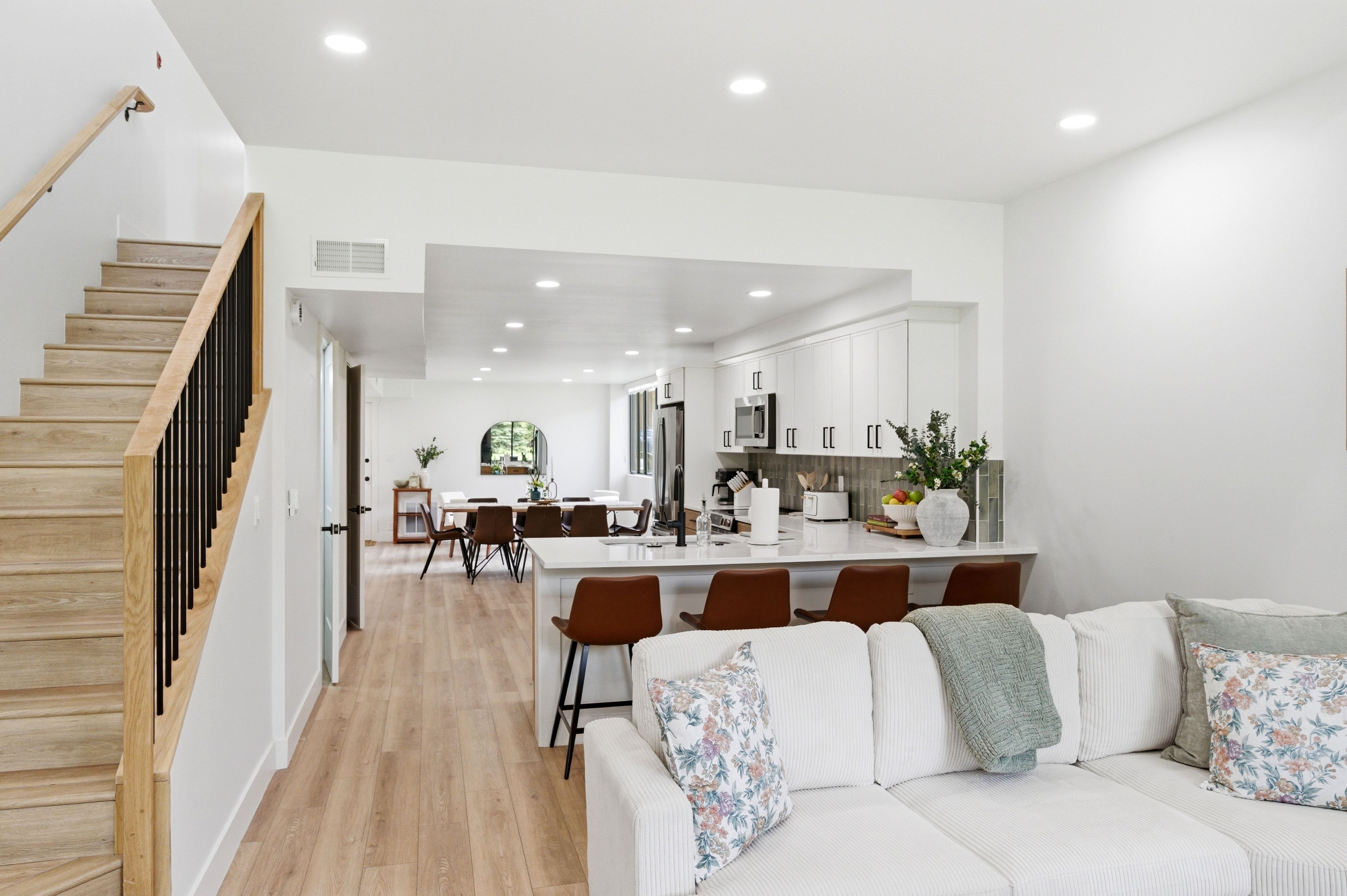 Open-concept living space featuring wood-finish flooring, a contemporary staircase with natural wood handrail and black metal balusters, recessed lighting, white cabinetry, and a kitchen island with seating