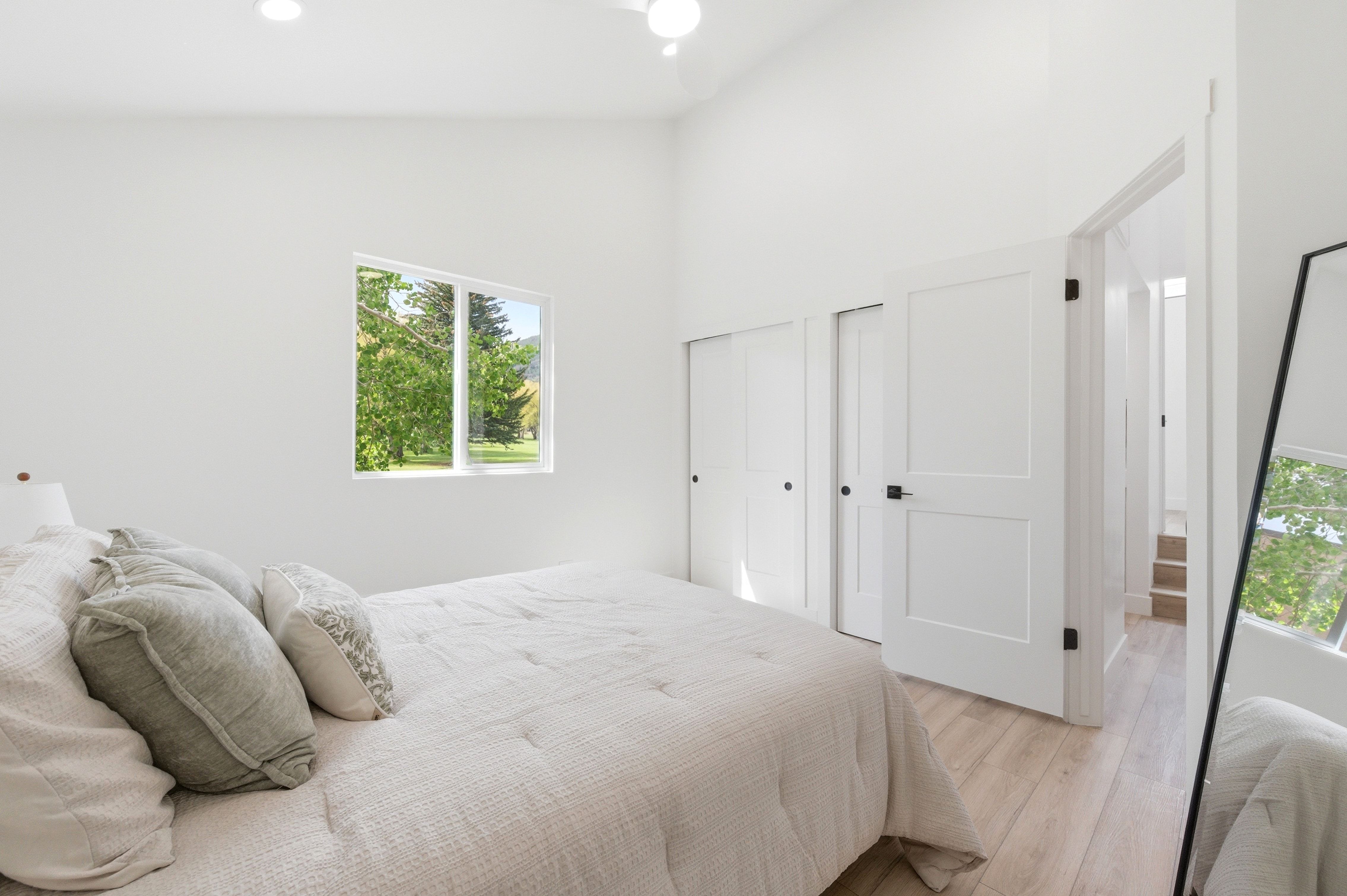 Light-filled bedroom featuring a window with green outdoor scenery, vaulted ceilings, recessed lighting, wood-finish flooring, and two panel doors with matte black hardware
