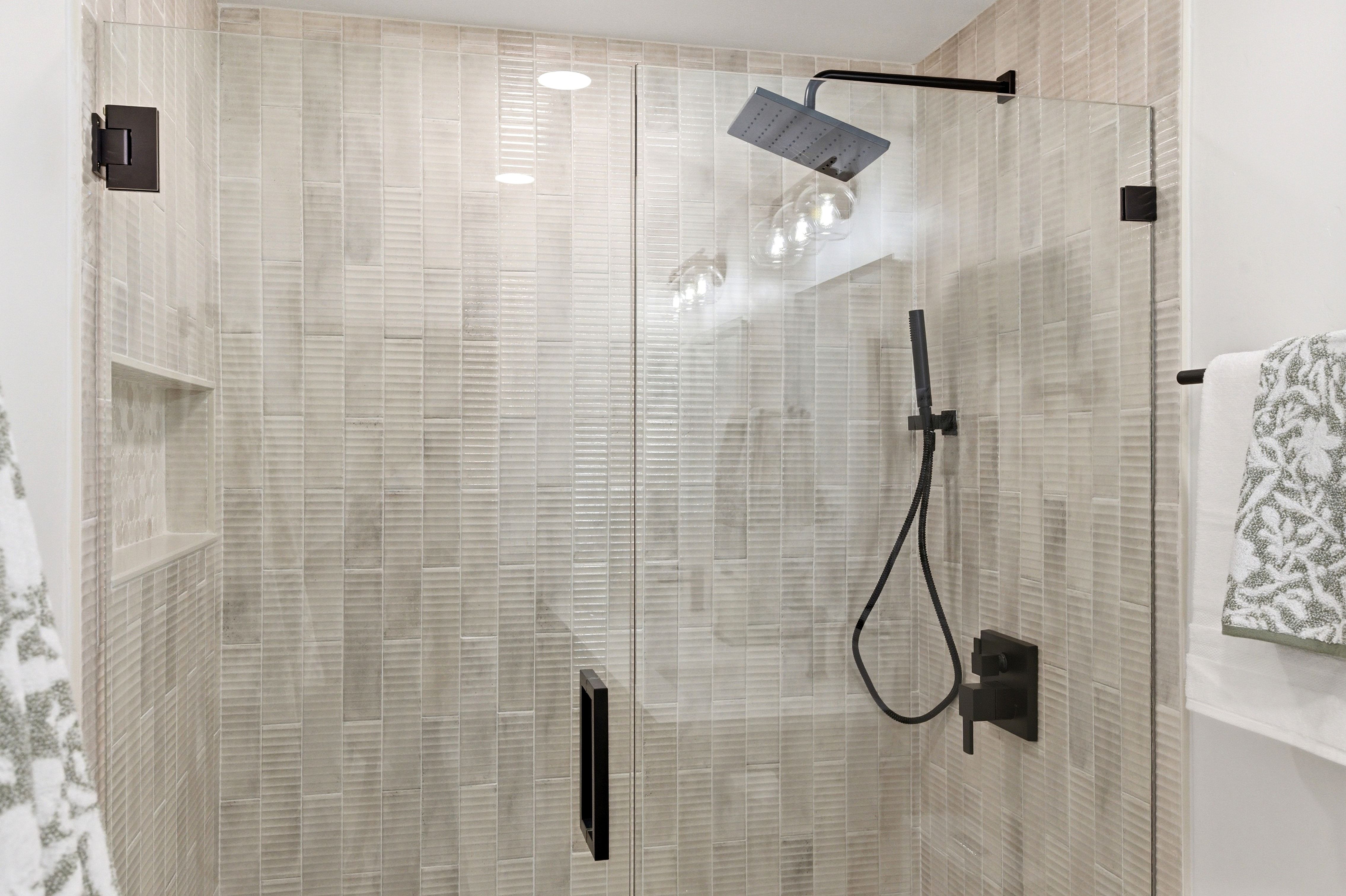 Glass-enclosed shower featuring vertically stacked tile, a built-in shower niche, and matte black fixtures including a rainfall shower head and handheld sprayer