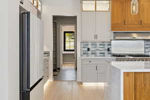 Gourmet kitchen featuring a professional-grade range with a stainless steel hood, light wood-finish flooring, a contemporary mosaic tile backsplash, custom cabinetry with integrated lighting, and a waterfall island with a light-toned countertop