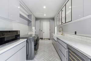 Galley-style utility room featuring light gray cabinetry, white countertops, herringbone-patterned floor tile, a built-in wine cooler, and a farmhouse-style sink with a gold-tone faucet