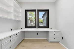 Dedicated office space featuring two windows with black frames, built-in shelving, light wood-finish flooring, and a wrap-around desk with white countertops and light gray cabinetry