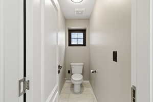 Compact powder room featuring a toilet, a small window with dark trim, light-colored wall paint, tiled flooring, and a ceiling light fixture