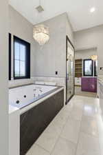 Soaking tub with a dark stone-look surround, white ceramic tile, and a decorative crystal chandelier