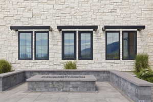 Stone exterior with dark-framed windows under black awnings, complemented by a paver patio featuring a built-in fire pit and retaining wall seating