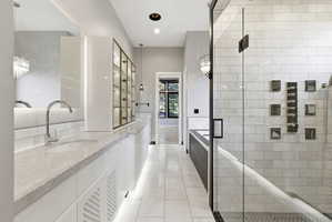 Spacious bathroom featuring dual vanity with undermount sinks and chrome fixtures, white tile flooring, a glass-enclosed shower with subway tile surround, and built-in display cabinetry