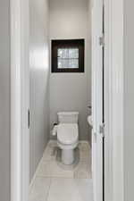 Powder room featuring a square window with dark trim, light-toned tile flooring, and a white toilet