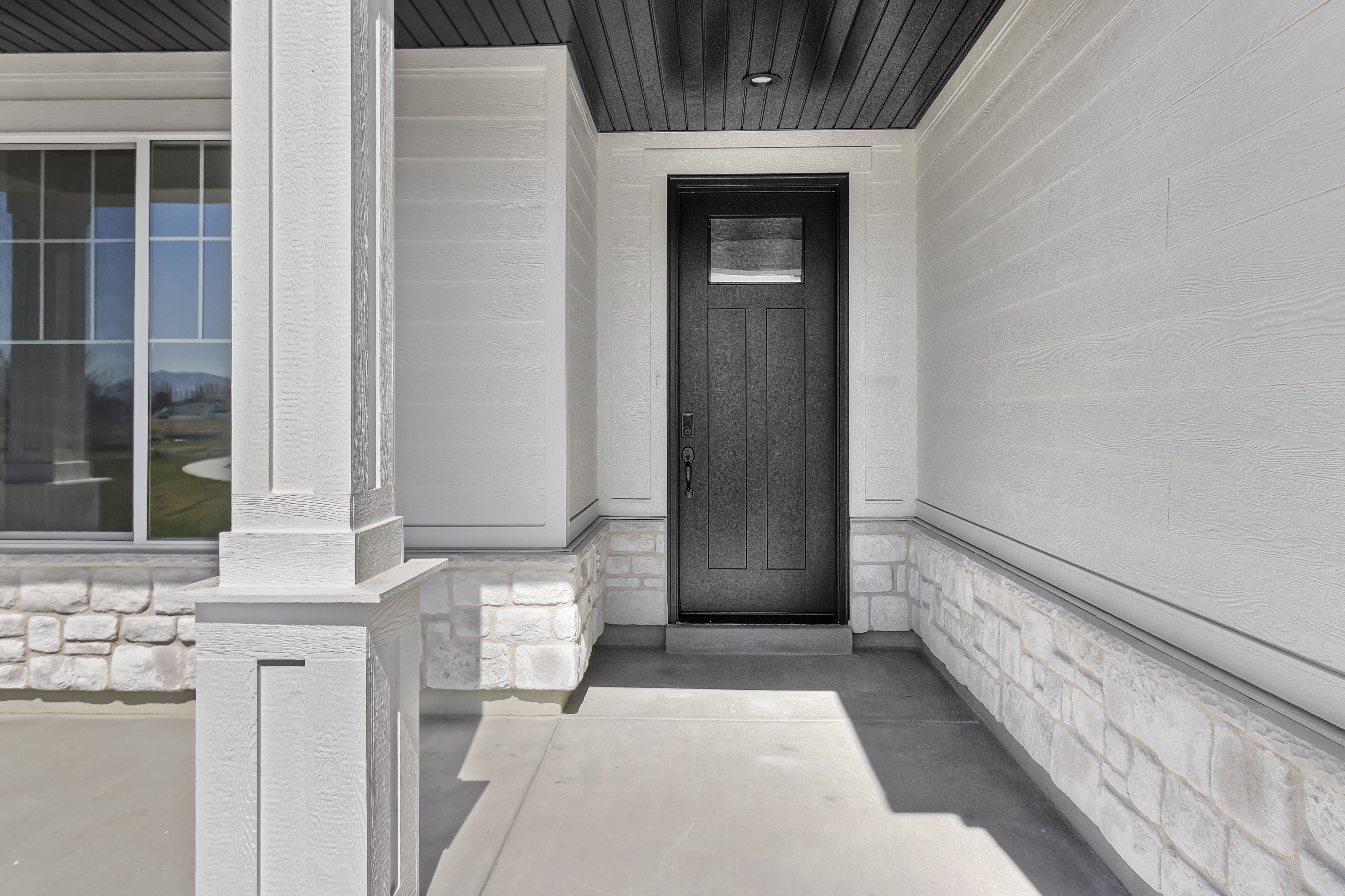 Contemporary entryway featuring a black panel door with transom window, recessed lighting, white siding, and a stone veneer foundation