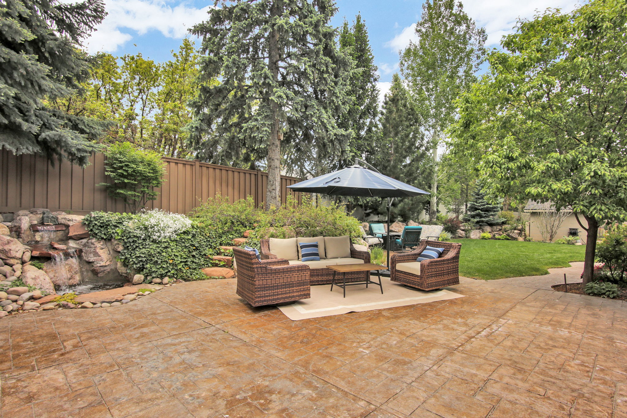 Expansive stamped concrete patio featuring a rock waterfall, mature evergreen trees, a manicured lawn, and a privacy fence