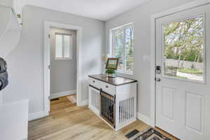 Entryway featuring a white paneled door with a glass insert, wood-finish flooring, light grey walls, and a built-in white storage bench with brass hardware