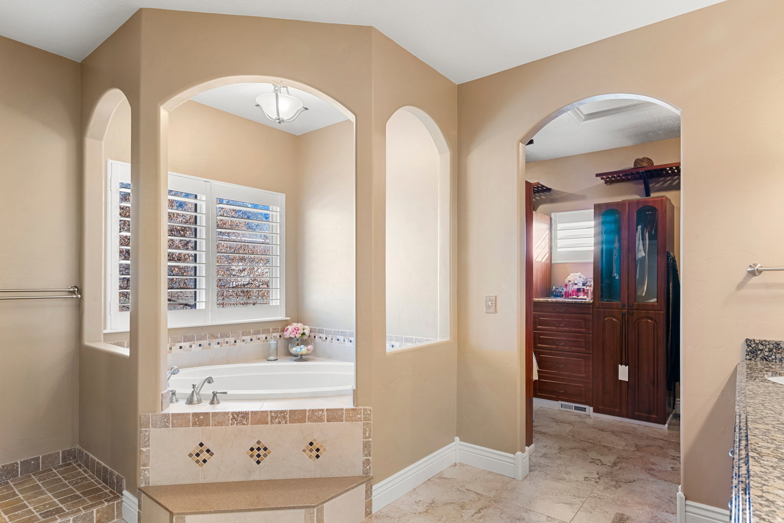 Recessed soaking tub with surrounding decorative tile accents, plantation shutters, and overhead lighting