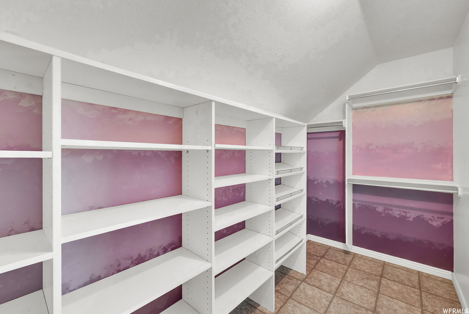 Spacious storage area featuring multiple white shelving units, integrated sliding wire baskets, ceramic tile flooring, and sloped ceiling details