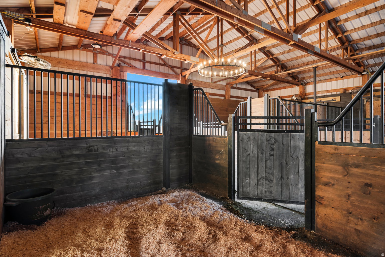 Barn interior featuring multiple stalls with wood and black metal construction, exposed timber ceiling beams, metal roofing, and a wagon wheel chandelier