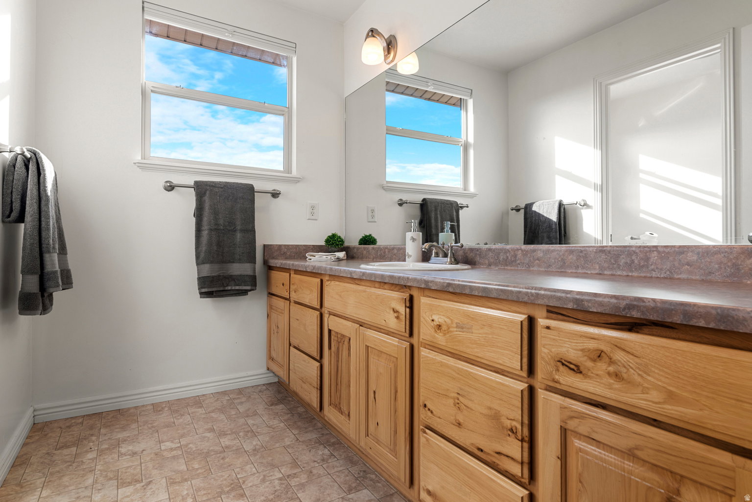 Dual windows with white blinds, a full-width vanity mirror, a double vanity with wood cabinetry, a built-in sink, and patterned floor tiling