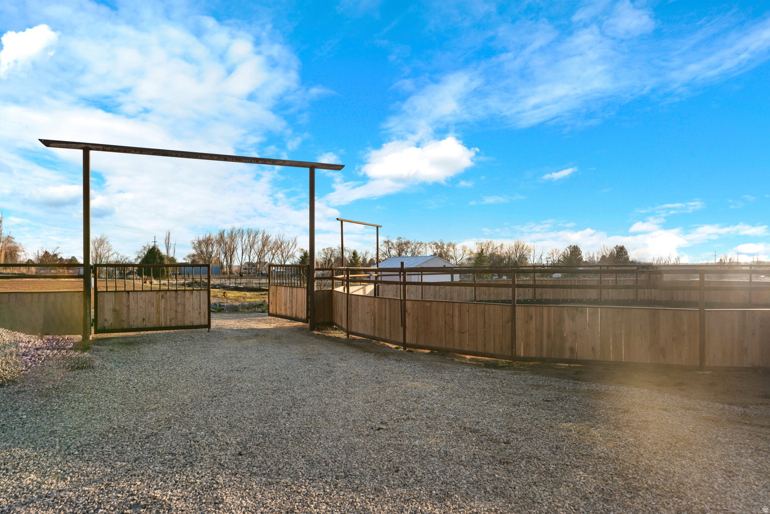Gravel drive entry with metal archway, leading to a fenced outdoor arena featuring wood-panel and metal-rail construction