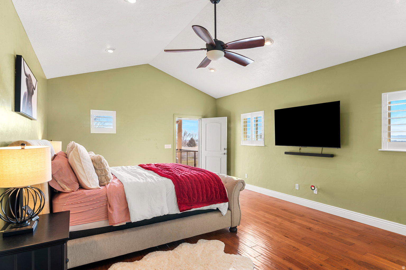 Spacious room featuring vaulted ceilings, wood-finish flooring, a ceiling fan, and multiple windows with plantation shutters