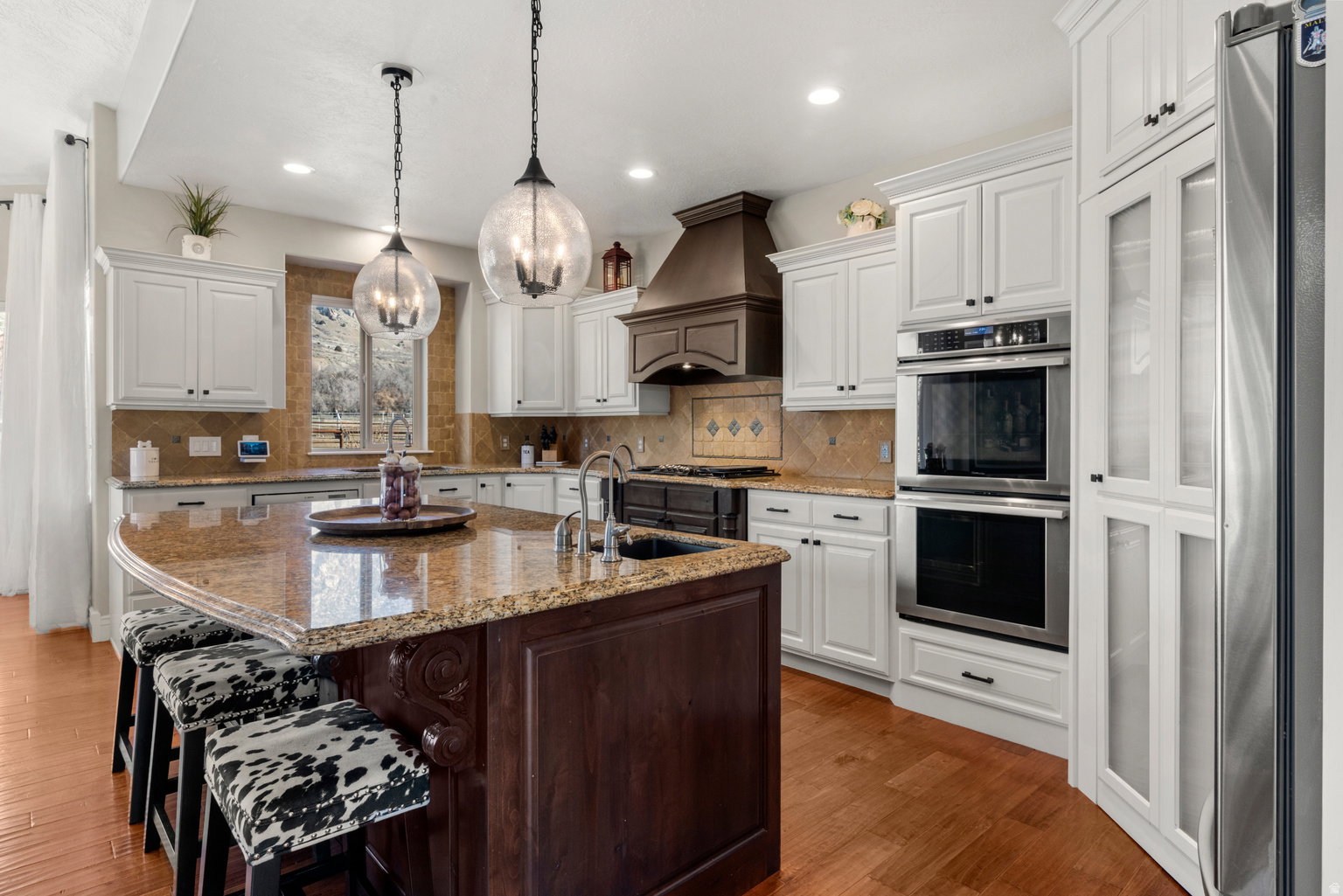 Kitchen featuring a substantial island with a granite countertop, white perimeter cabinetry, stainless steel double wall ovens, a dark wood-finish range hood, and wood-finish flooring
