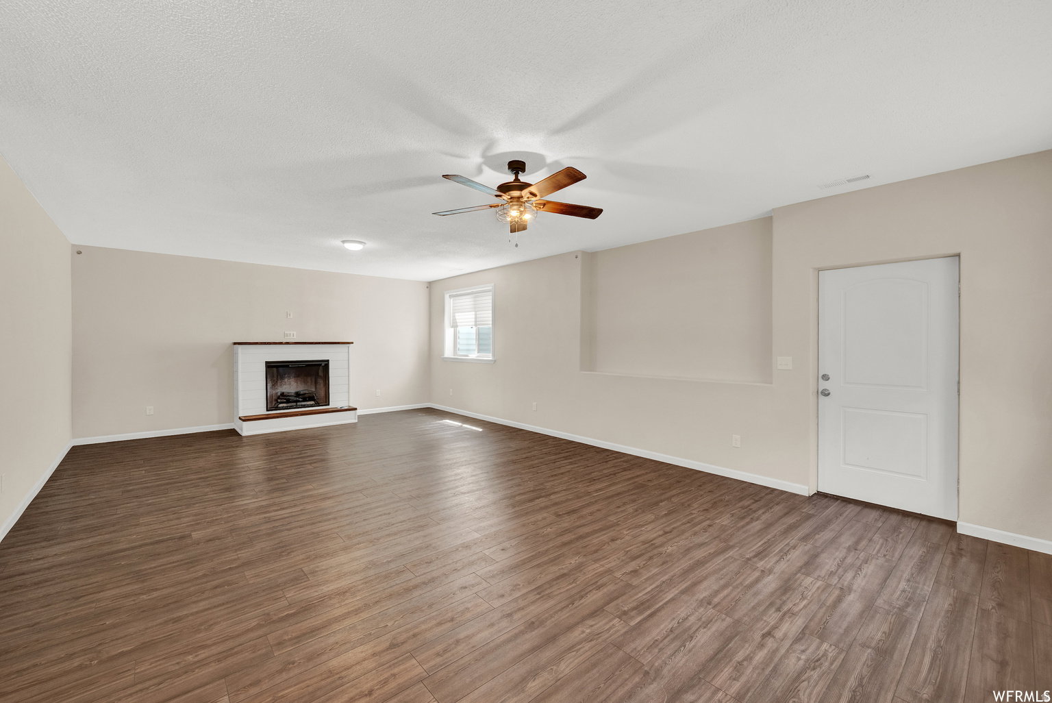 Spacious room featuring wood-finish flooring, a white brick fireplace with a wood mantel, and a ceiling fan with integrated lighting