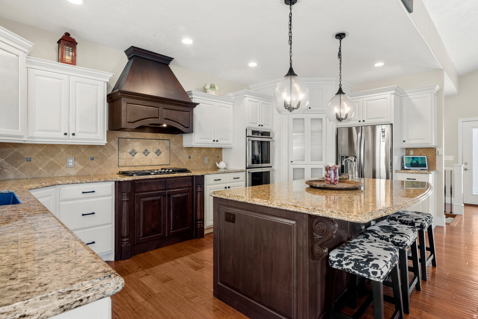Kitchen featuring a wood-finish floor, light-toned granite countertops, white cabinetry with dark hardware, a dark wood-finish range hood, and a stainless steel refrigerator