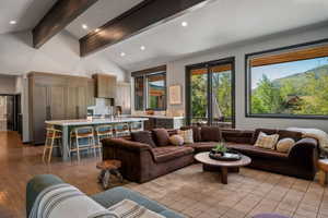 Open-concept living area featuring vaulted ceilings with exposed beams, wood-finish flooring, a kitchen island with seating, integrated appliances, and large windows with mountain scenery