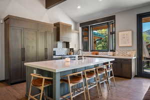 Kitchen featuring a large central island with seating, a paneled refrigerator, wood-finish flooring, a vaulted ceiling, and a geometric tile backsplash