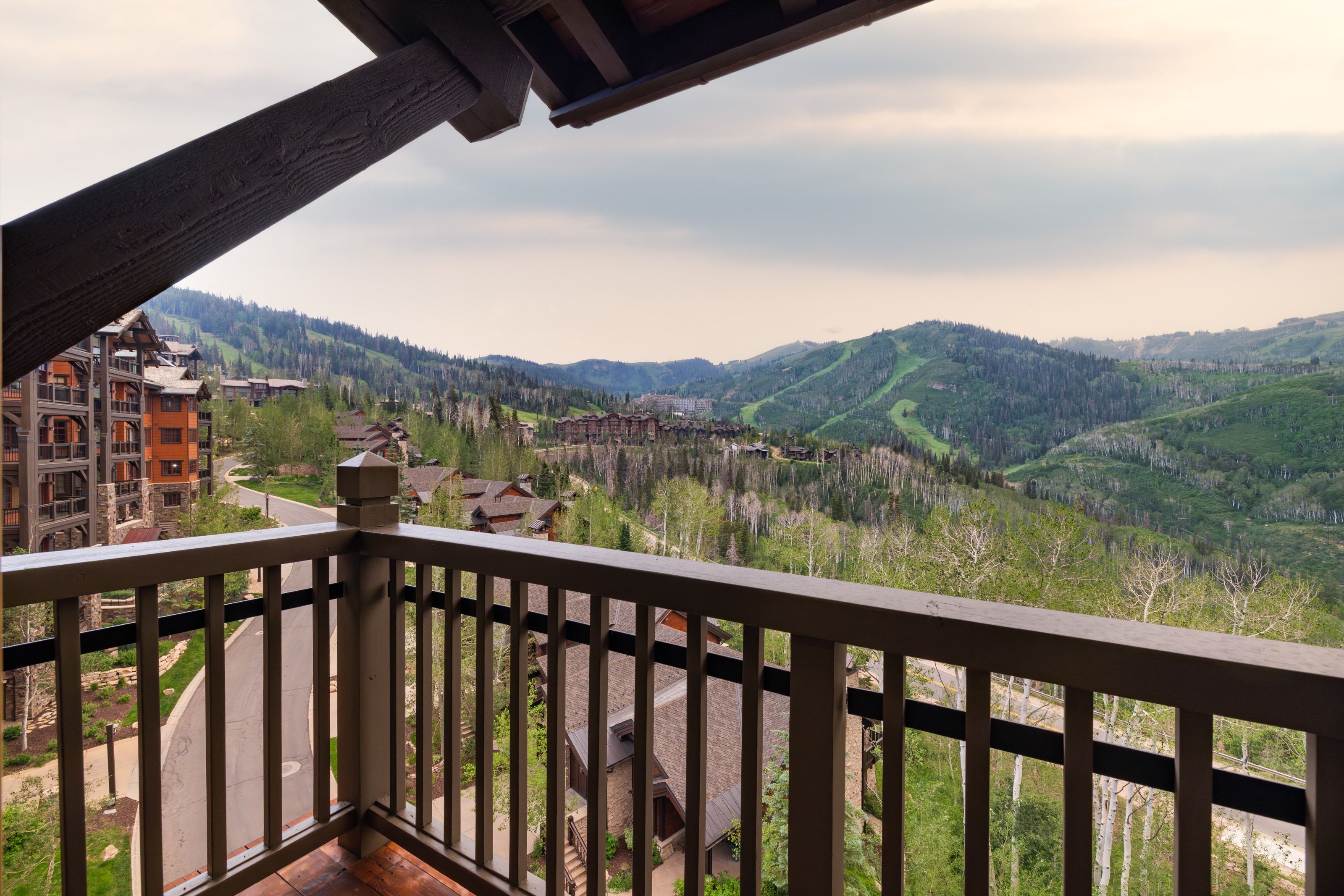 Balcony with wood railing and supporting beam, offering expansive mountainous terrain with evergreen and deciduous trees, and distant structures